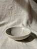 A small handmade ceramic bowl for sage and pale santo with a speckled off-white glaze, resting on draped beige linen fabric under natural sunlight.