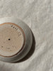 A close-up of the bottom of a round ceramic dish, showing a speckled beige and brown glaze with a stamped maker's mark and number, placed on a textured off-white fabric surface. The makers mark spells 'sati'