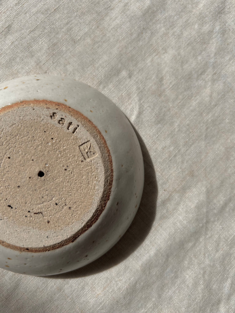 A close-up of the bottom of a round ceramic dish, showing a speckled beige and brown glaze with a stamped maker's mark and number, placed on a textured off-white fabric surface. The makers mark spells 'sati'