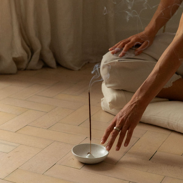 A person kneeling on a light wood floor beside a white ceramic incense holder with a smoking incense stick, in a softly lit indoor setting with neutral-toned cushions and draped fabric in the background.