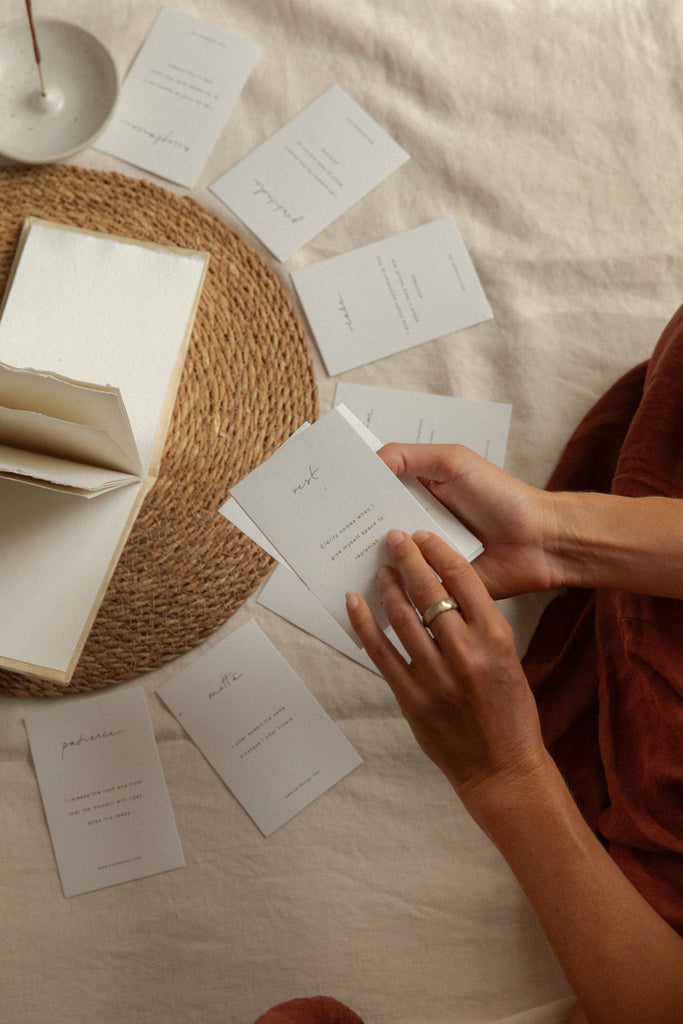 A top-down view of a person's hands holding some affirmation cards on a beige linen surface, surrounded by an open book, a woven placemat, and other affirmation cards.