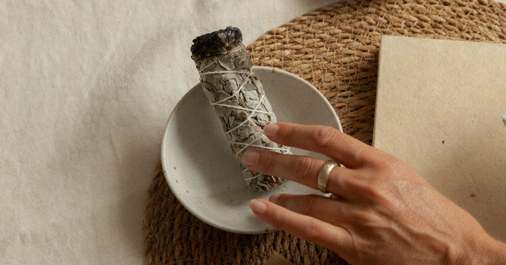 A hand holds a smudge bundle wrapped in white sage over a small white dish, with an incense holder, paper, and kraft paper boxes on woven placemats. The brand name 'sati' is visible on the box.