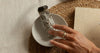A hand holds a smudge bundle wrapped in white sage over a small white dish, with an incense holder, paper, and kraft paper boxes on woven placemats. The brand name 'sati' is visible on the box.