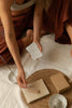 A person in rust-coloured trousers sits on a white fabric floor, holding a small white affirmation card over a set of incense-related items including a woven tray, a beige book, a small white dish with a incense stick, and a kraft paper box.
