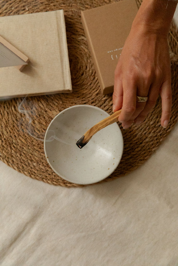A hand holds a burning palo santo stick over a small speckled white ceramic bowl, placed on a woven jute placemat with neutral-toned books in the background.
