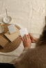 A person holds a small white affirmation card over a woven mat on a cream fabric surface, with more affirmation cards, a kraft box and a thin incense stick in a ceramic dish nearby.