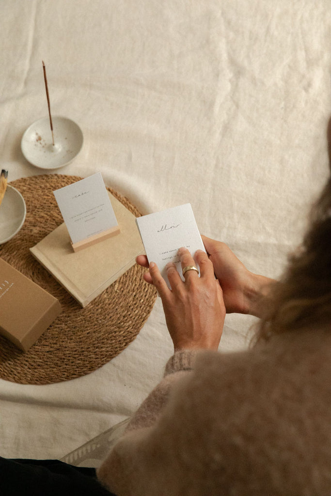 A person holds a small white affirmation card over a woven mat on a cream fabric surface, with more affirmation cards, a kraft box and a thin incense stick in a ceramic dish nearby.