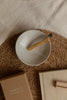 A top-down styled product shot of a small speckled off-white ceramic bowl with a palo santo stick resting inside, placed on a round woven jute mat over a beige fabric surface. A brown kraft box with the brand name '{sati}' is visible at the bottom left, and a beige fabric-covered book or box sits at the bottom right.