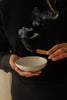 A person in a black sweater holds a small speckled white ceramic bowl while lighting a palo santo stick, with smoke rising above the bowl.