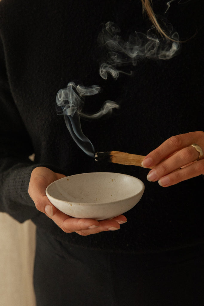 A person in a black sweater holds a small speckled white ceramic bowl while lighting a palo santo stick, with smoke rising above the bowl.