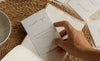 A hand holds a small white affirmation card over an open book on a woven placemat, with other affirmation cards and a ceramic incense holder on a neutral fabric background.