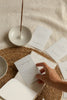 A hand holds a small white affirmation card over an open book on a woven placemat, with other affirmation cards and a ceramic incense holder on a neutral fabric background.