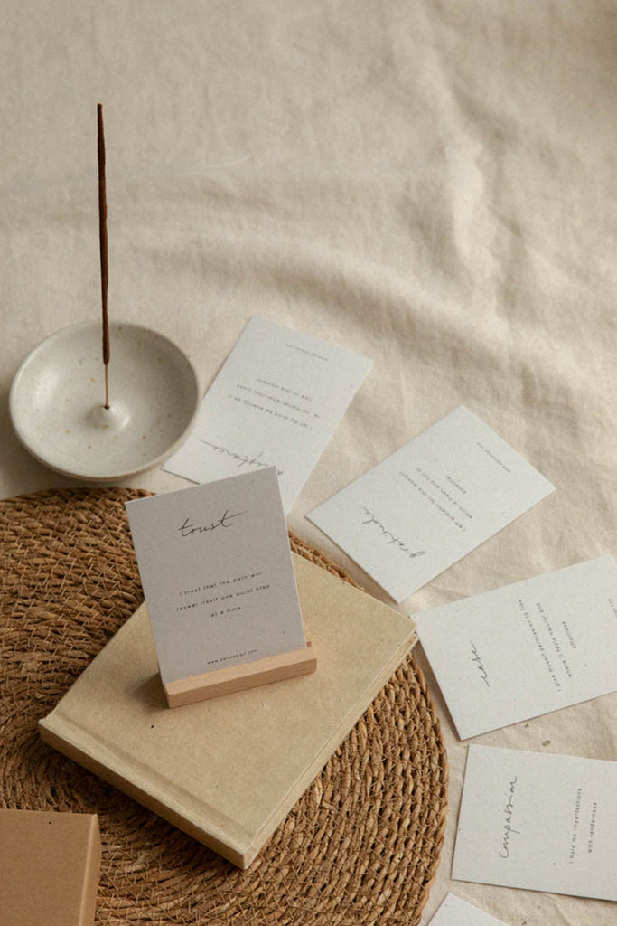 Minimal affirmation cards arranged on a woven jute placemat with a beige linen backdrop. A small white dish with an incense stick sits at left, and several white affirmation cards with delicate black script and block text are spread out on a beige book.