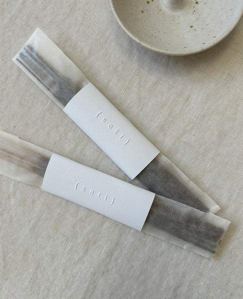 Packaged incense sticks wrapped in clear sleeves with white paper bands embossed with the brand name '(sati)', arranged on a light beige linen surface with a ceramic incense stick holder partially visible in the corner.