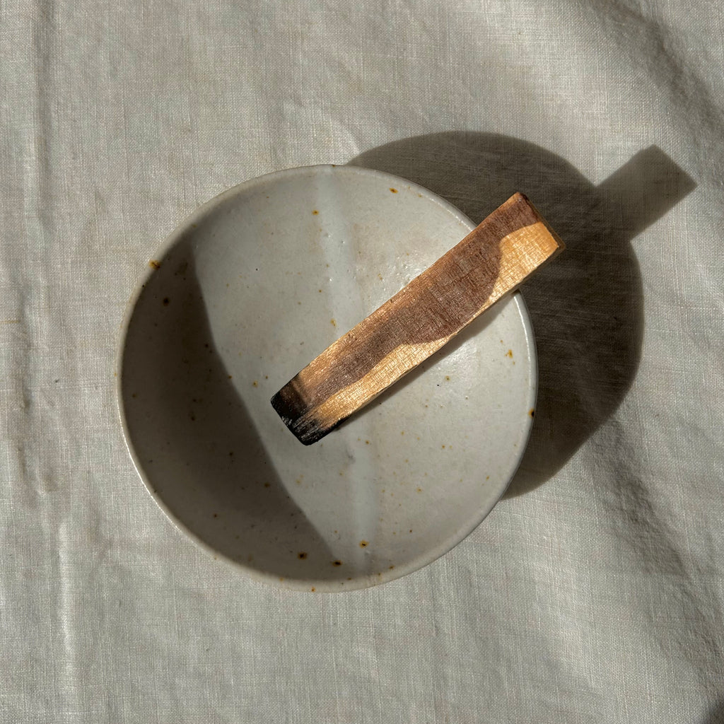 A handmade ceramic bowl with a speckled glaze sits on a textured linen cloth, with a stick of just-burned palo santo resting inside it under direct sunlight.