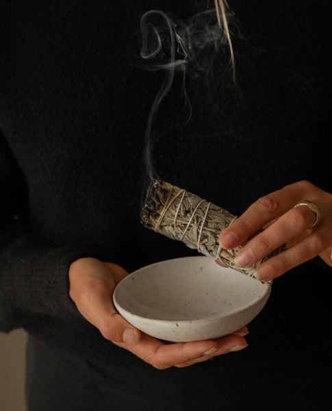 A person in a black sweater holds a sage smudge bundle with smoke rising above a small speckled ceramic bowl.